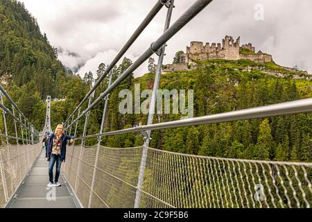 Angela Berg geht lächelnd über die Hochwurfen179-Fußgängerklappbrücke nahe Reutte, Tirol, Österreich, mit den Ruinen der Burg Ehrenberg im Hintergrund. Die 406 Meter lange Brücke verbindet die Burgruinen mit dem gegenüberliegenden Fort Claudia und überspannt das Tal in einer Höhe von bis zu 114 Metern; von ihrer Eröffnung im Jahr 2014 bis 2017 hielt sie den Guinness-Weltrekord als längste Fußgängerklappbrücke der Welt. Stockfoto