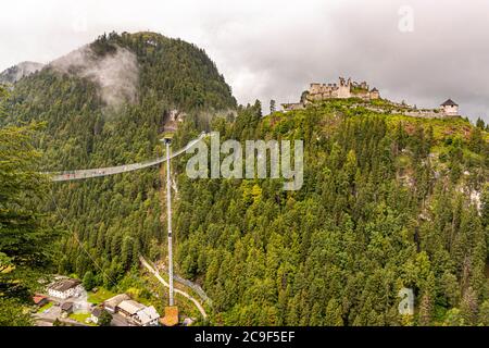 Leichte Nebelschwaden ziehen über den bewaldeten Hang unter den Ruinen der Burg Ehrenberg bei Reutte in Tirol, Österreich, während die Highline179-Fußgängerklammbrücke vom AnkerTurm im Tal zum Burganlage aufsteigt. Die 406 Meter lange Brücke überspannt das Tal in einer Höhe von bis zu 114 Metern und verbindet die Burgruine mit dem gegenüberliegenden Fort Claudia; von ihrer Eröffnung im Jahr 2014 bis 2017 hielt sie den Guinness-Weltrekord als längste Fußgängerklammbrücke der Welt. Stockfoto