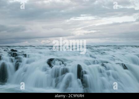 Ein großer Wasserkörper fließt über dunkle Felsen an der Spitze eines Wasserfalls. Das Wasser ist blau/weiß und kalt, ähnlich wie die Wolken oben am Himmel. Stockfoto