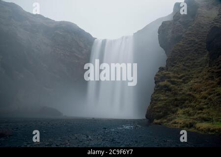 Ein Wasserfall fließt vor dunklen, moosigen Klippen hinunter. Nebel driftet über die schwarzen Felsen darunter. Der Himmel ist hellgrau. Stockfoto