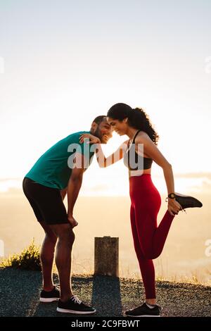 Fröhliches Paar, das morgens auf der Straße steht und sich aufwärmt. Porträt einer athletischen Frau, die ihre Beinmuskeln dehnt. Stockfoto