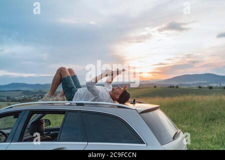 Der junge Mann lag auf dem Autodach und las das Papier-Bestseller-Buch.Er hielt sein Auto auf der Wiese mit einem schönen Blick auf das Tal vor dem Sonnenuntergang an Stockfoto