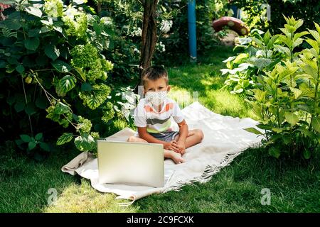 Kleiner Junge in einer Maske, der während der Quarantänezeit auf dem Gras mit einem Laptop auf Plaid liegt. Stockfoto