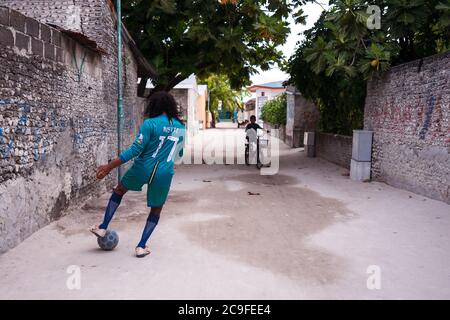 Bodufolhudhoo / Malediven - 17. August 2019: Junger maledivischer Mann mit langen lockigen Haaren spielt mit Fußball auf der Straße der Inselstadt Stockfoto