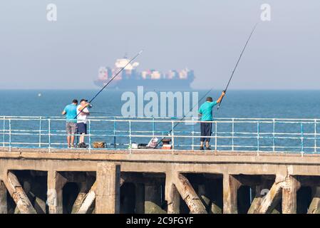 Angler Angeln vor Barge Pier in der Themse Mündung vor Shoeburyness, Southend on Sea, Essex, Großbritannien, mit großen Frachtschiff vorbei Stockfoto