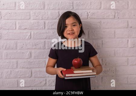 Zurück zur Schule. Kleines Mädchen hält Bücher und einen Apfel in der Nähe einer weißen Ziegelmauer. Kind aus der Grundschule. Stockfoto