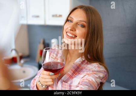 Glückliche, entspannte junge Frau, die mit einem Glas Rotwein in der Küche steht und ihr Smartphone benutzt Stockfoto