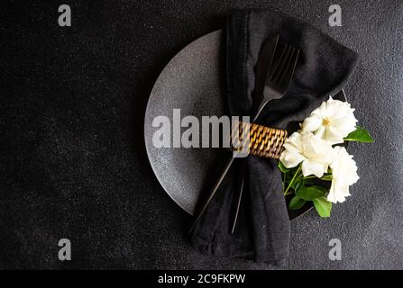 Minimalistisches Tischdekor mit schwarzem Steinzeug und Besteck mit weißer Rosenblüte auf schwarzem Steintisch mit Kopierfläche Stockfoto