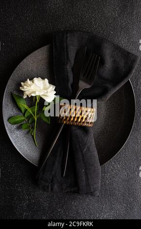 Minimalistisches Tischdekor mit schwarzem Steinzeug und Besteck mit weißer Rosenblüte auf schwarzem Steintisch mit Kopierfläche Stockfoto