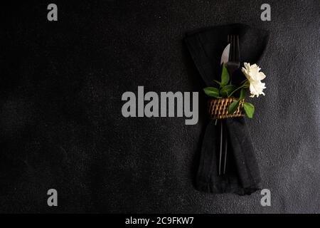 Minimalistisches Tischdekor mit schwarzem Steinzeug und Besteck mit weißer Rosenblüte auf schwarzem Steintisch mit Kopierfläche Stockfoto