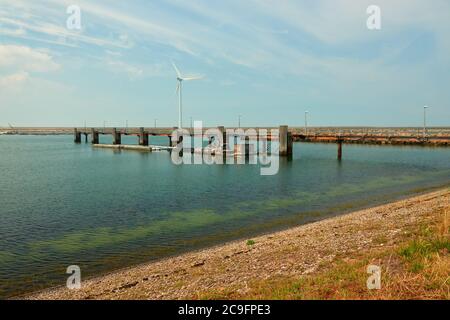 Brauner Kiesstrand und tiefblaues Wasser, ein langer Pier mit Holzsäulen, Windturbine und Damm im Hintergrund. Niederlande, Vrowenpolder. Stockfoto