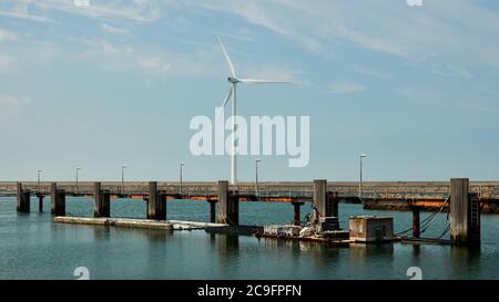 Langer Steg aus Stahl und Holz führt über das Wasser, große Windturbine auf dem Damm. Niederlande, Vrouwenpolder. Stockfoto