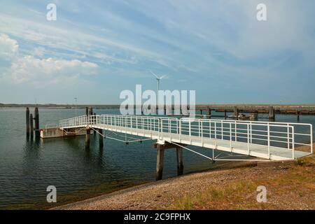 Der lange weiße Pier führt zum Bootsanleger. Windturbine und Damm im Hintergrund. Zeeland, Niederlande. Stockfoto