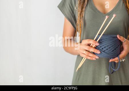 Eine junge Strickerin mit Dreadlocks in einem grünen Kleid hält einen Knäuel aus blauem Garn und Holzstricknadeln zum Stricken. Heller Hintergrund. Stockfoto