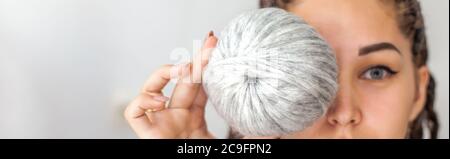 Eine schöne junge Strickerin lächelt und hält Skeins aus hellgrauem und weißem Garn in den Händen. Frau mit Dreadlocks. Stockfoto