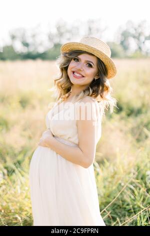 Lächelnd charmante junge schwangere Frau, trägt weißes Kleid und Strohhut, Ruhe im Sommerfeld. Im Freien posieren. Mutterschaft. Außenportrait von Stockfoto