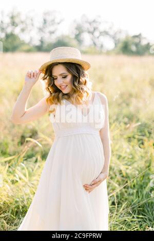 Lächelnd charmante junge schwangere Frau, trägt weißes Kleid und Strohhut, Ruhe im Sommerfeld. Im Freien posieren. Mutterschaft. Außenportrait von Stockfoto