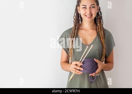 Eine junge Strickerin mit Dreadlocks in einem grünen Kleid hält einen Knäuel aus blauem Garn und Holzstricknadeln zum Stricken. Heller Hintergrund. Stockfoto