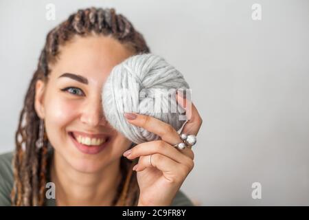 Eine schöne junge Strickerin lächelt und hält Skeins aus hellgrauem und weißem Garn in den Händen. Frau mit Dreadlocks. Stockfoto