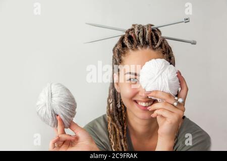 Eine schöne junge Strickerin lächelt und hält Skeins aus hellgrauem und weißem Garn in den Händen. Frau mit Dreadlocks. Stockfoto