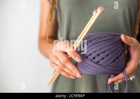 Eine junge Strickerin mit Dreadlocks in einem grünen Kleid hält einen Knäuel aus violettem Garn und Holzstricknadeln zum Stricken. Heller Hintergrund. Stockfoto