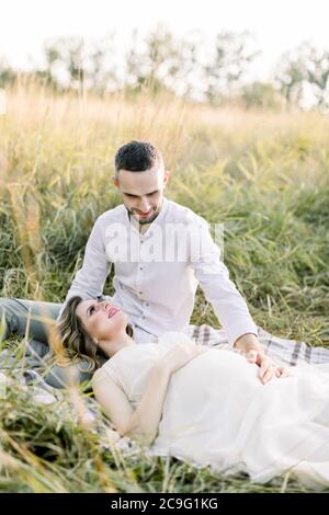 Schöner bärtiger kaukasischer Mann, der seine charmante schwangere Frau ansieht, die sich in warmen Sommerabenden auf einer Decke auf dem Gras auf dem Feld ausruht Stockfoto