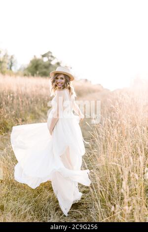 Lächelnd charmante junge schwangere Frau, trägt weißes Kleid und Strohhut, Ruhe im Sommerfeld. Im Freien posieren. Mutterschaft. Außenportrait von Stockfoto