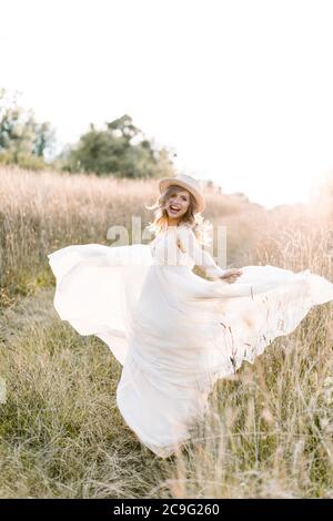 Lächelnd charmante junge schwangere Frau, trägt weißes Kleid und Strohhut, Ruhe im Sommerfeld. Im Freien posieren. Mutterschaft. Außenportrait von Stockfoto