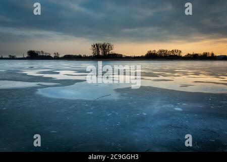 Wasserpfützen auf dem gefrorenen See und Wolken nach Sonnenuntergang, Winterabendlandschaft Stockfoto