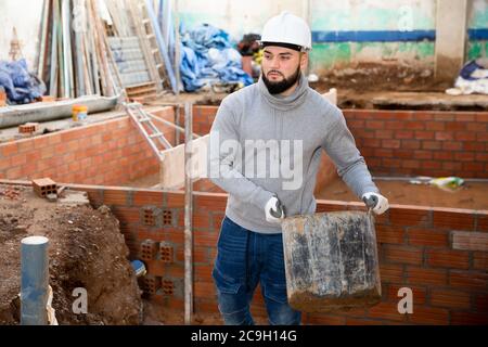 Selbstbewusster bärtiger Kerl, der auf der Baustelle arbeitet und Eimer Baumörtel trägt Stockfoto