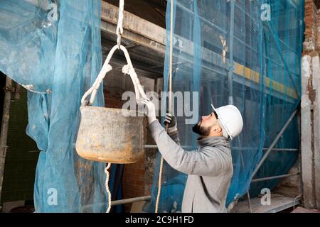 Junger bärtiger Mann, der auf der Baustelle arbeitet und Eimer mit Baumörtel am Seil hebt Stockfoto