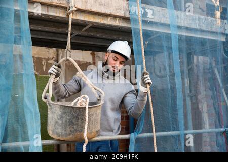 Selbstbewusster bärtiger Kerl im Bauhelm, der an seiner Hausrenovierung arbeitet und mit Werkzeugen am Seil den Eimer heruntersenkt Stockfoto