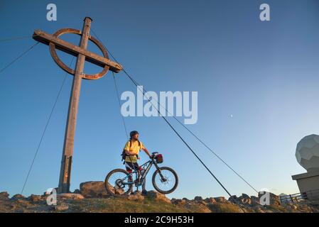 Mountainbiker steht mit E-MTB neben dem Gipfelkreuz des Patscherkofels im warmen Abendlicht, Patscherkofel, Patsch, Tirol, Österreich Stockfoto