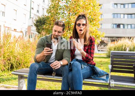 Ein Paar ist schockiert über Inhalte auf dem Bildschirm eines Telefons gesehen, während im Sommer auf einer Parkbank in der Stadt sitzen. Stockfoto