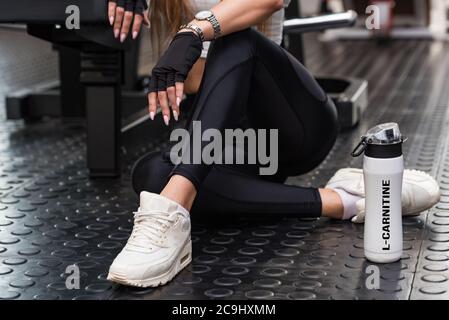 Attraktives Modell sitzt mit L-Carnitin Drink in einem modernen Fitnessraum. Unteren Teil anzeigen Stockfoto