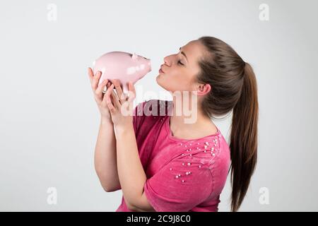Junge schöne Frau mit Ponitail küsst ein Sparschwein, liebt Geld, gegen rosa Hinterlauf Stockfoto
