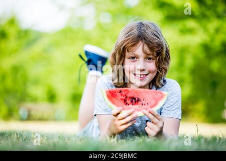 Junge hält Wassermelone, während er im Park liegt, lächelt, Porträt. Stockfoto