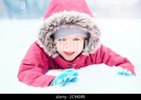 Cute kaukasischen lächelnd aufgeregt Mädchen Kind in rosa Jacke mit Schnee spielen. Kind liegt auf dem Boden während des kalten Winterschneetags bei Schneefall. Kinder im Freien Stockfoto