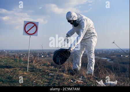 Wissenschaftler tragen weiße Schutzhülle, Gasmaske, sammeln Plastikmüll in schwarzen Abfallbeutel im Freien an einem sonnigen Tag im Frühjahr, unter blauem Himmel, neben Verbotssymbol, Reinigung Gebiet Stockfoto