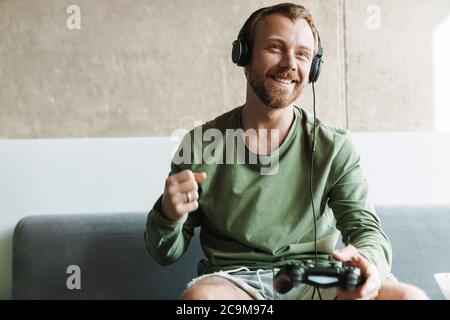 Foto des lachenden Mannes, der Kopfhörer trägt und Videospiele spielt, während er zu Hause auf dem Sofa eine Gewinnergeste macht Stockfoto