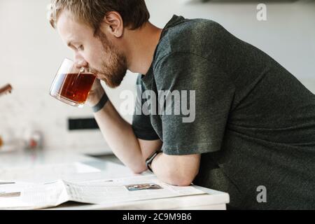 Foto eines ernsthaften Rotschopf-Mannes mit Bart Zeitung lesen, während Tee in der Küche zu Hause trinken Stockfoto