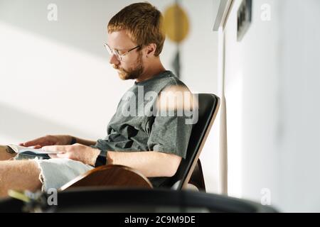 Foto des denkenden jungen Mannes, der Brillen trägt und Buch liest, während er zu Hause im Sessel sitzt Stockfoto