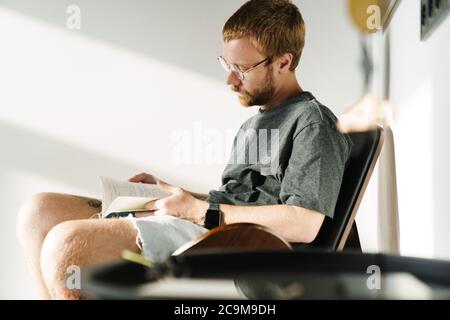Foto des denkenden jungen Mannes, der Brillen trägt und Buch liest, während er zu Hause im Sessel sitzt Stockfoto