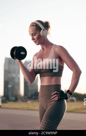 Junge Frau mit Hantel im Park. Sportliche Mädchen in Fitness trägt dabei körperliche Bewegung und Musik hören in Kopfhörer. Stockfoto