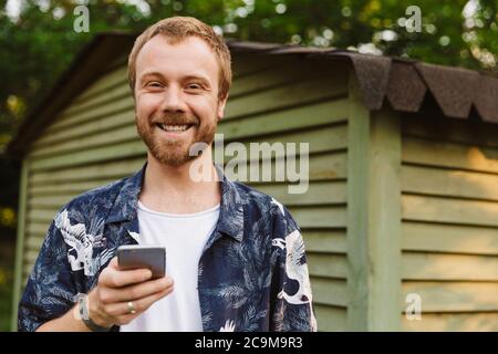Bild eines jungen kaukasischen lächelnden Mannes, der im Freien neben einem Holzhaus ein Handy hält Stockfoto