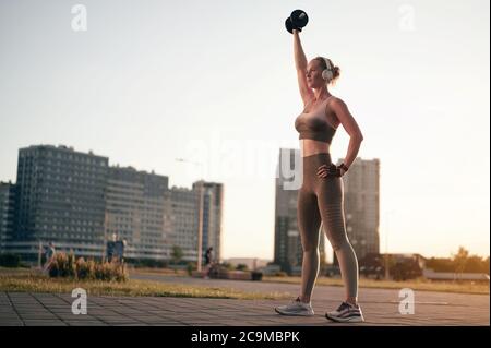 Fitness-Mädchen tun Übung mit Hantel im Freien. Sportlich starke Frau, die Gewicht verliert und Musik in Kopfhörern hört. Blick auf die Stadt, hintergrundbeleuchteter Sonnenuntergang Stockfoto