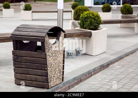 Ein mit Stein und Holzbohlen verzierter Mülleimer im Freien steht auf einem Gehweg aus grauen Fliesen in der Nähe von Holzbänken mit Blumentöpfen für Büsche. Stockfoto