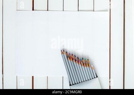 Mehrere Buntstifte, die aus einer Packung schauen, die auf dem Blatt Papier liegen, das auf einem Holztisch aus weißem Brett liegt. Stockfoto