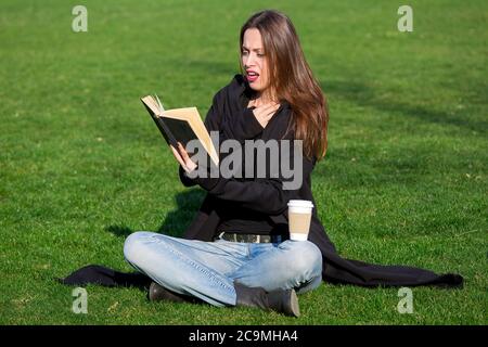 Mädchen mit Emotionen auf ihrem Gesicht liest ein Buch im Park sitzen auf dem grünen Gras an einem sonnigen Tag mit einer Einweg-Tasse Kaffee. Stockfoto