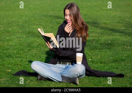 Ein junges Mädchen in einem schwarzen Mantel mit einem fröhlichen Lächeln auf ihrem Gesicht sitzt auf dem grünen Gras im Park und liest ein schwarzes Hardback-Buch und hält ihre Hand an Stockfoto